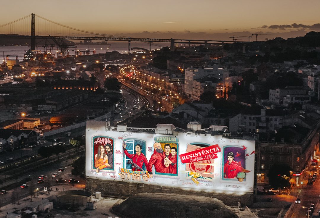 An expansive aerial night view of the massive "La Casa de Papel" tribute mural on Avenida 24 de Julho, framed by the Lisbon cityscape and the illuminated 25 de Abril Bridge, curated and produced by Because Art Matters in collaboration with Netflix.